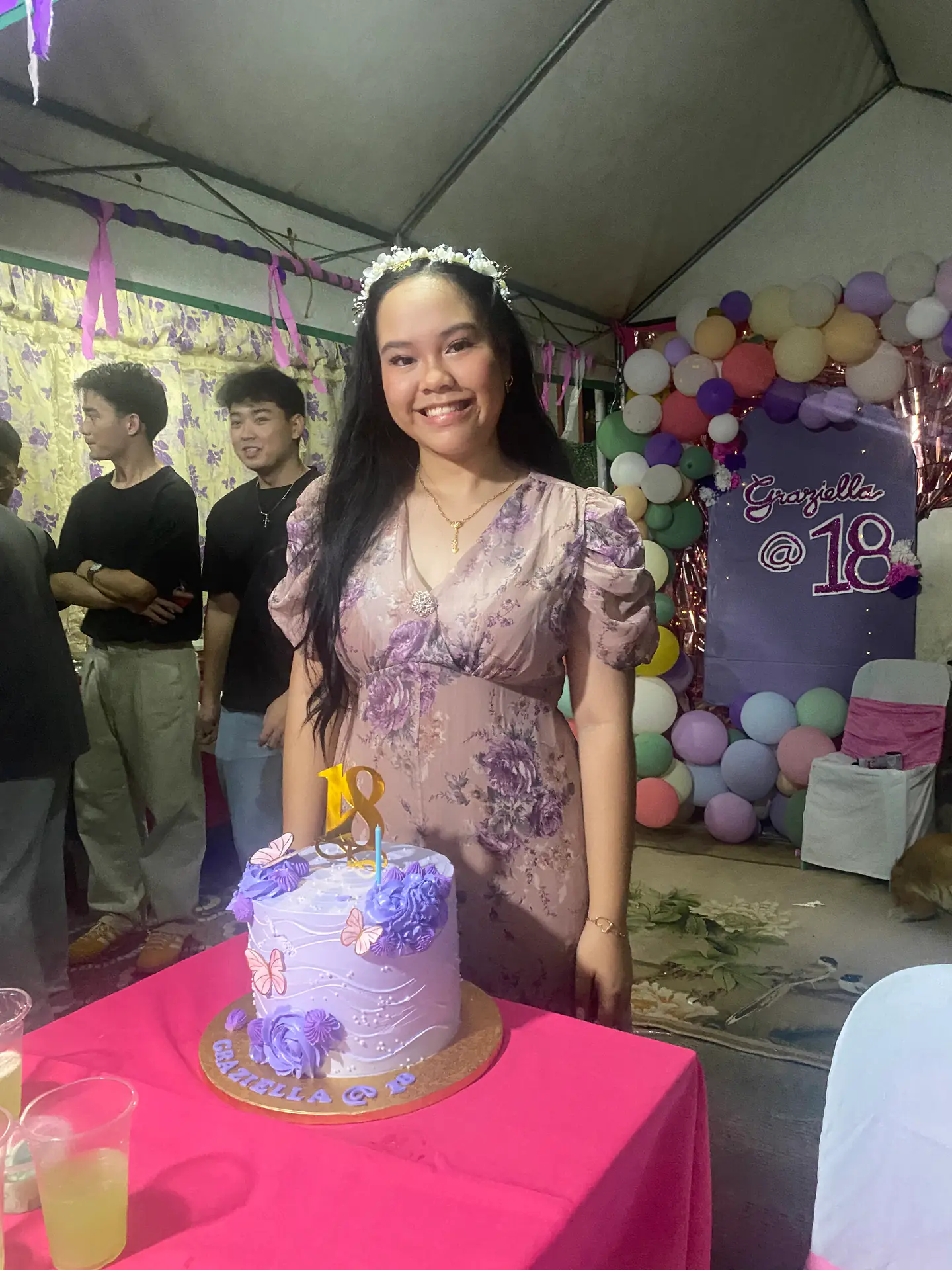A debut celebrant smiles beside a purple cake and pastel balloon backdrop.