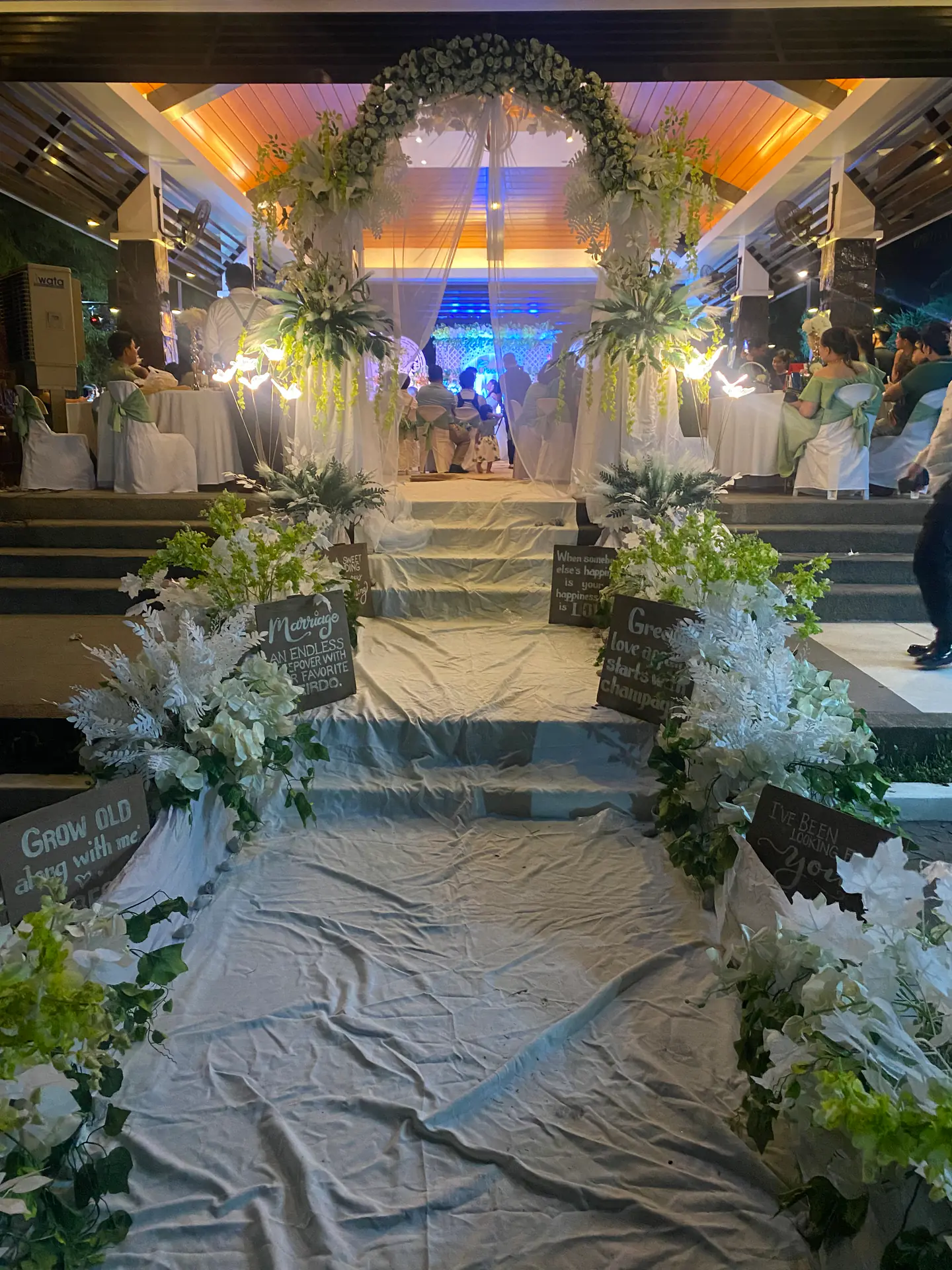 A wedding aisle framed by white flowers, greenery, soft fabric, and warm lights.